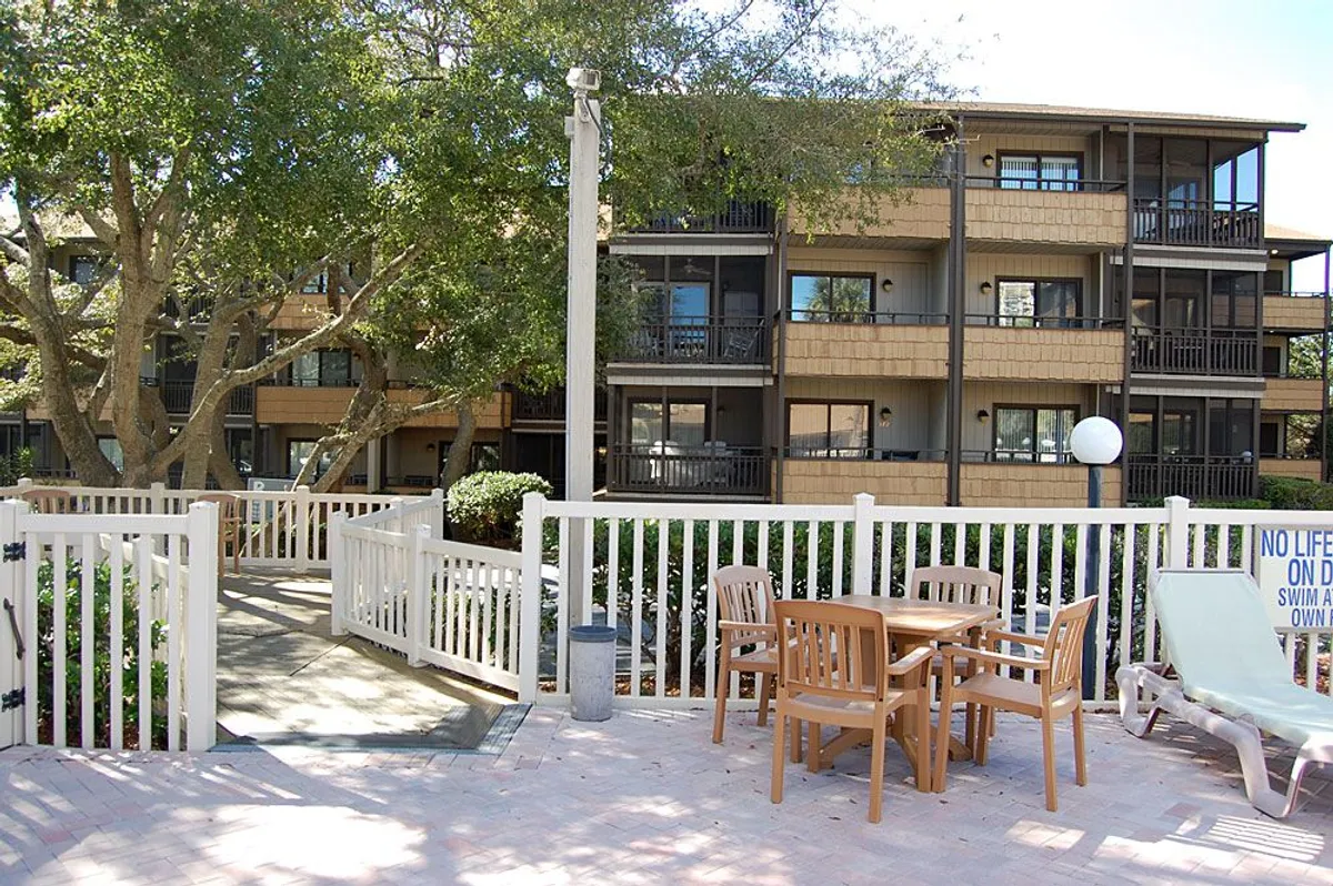 A comfortable outdoor seating area nestled beside the building facade, surrounded by lush greenery and shaded by trees. The space is inviting and designed for relaxation and socializing, with ample room for groups or quiet moments alone. The atmosphere suggests a perfect spot for morning coffee, afternoon chats, or evening relaxation, echoing the versatile use of the porch or balcony.