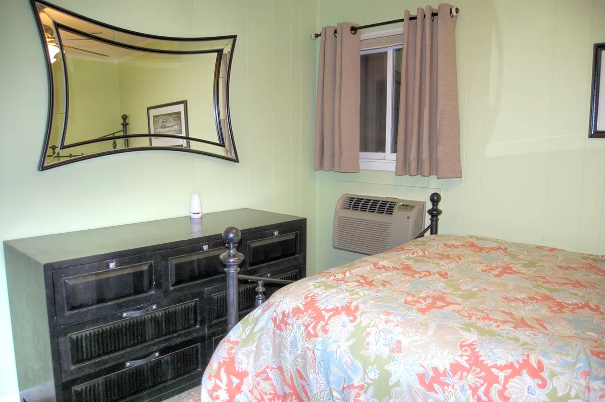 A cheerful bedroom featuring a bed with colorful bedding, a black dresser, mirror, and a window with curtains. The decor is lively yet inviting, and the natural light adds warmth to the setting, offering a cozy spot to unwind or start the day.
