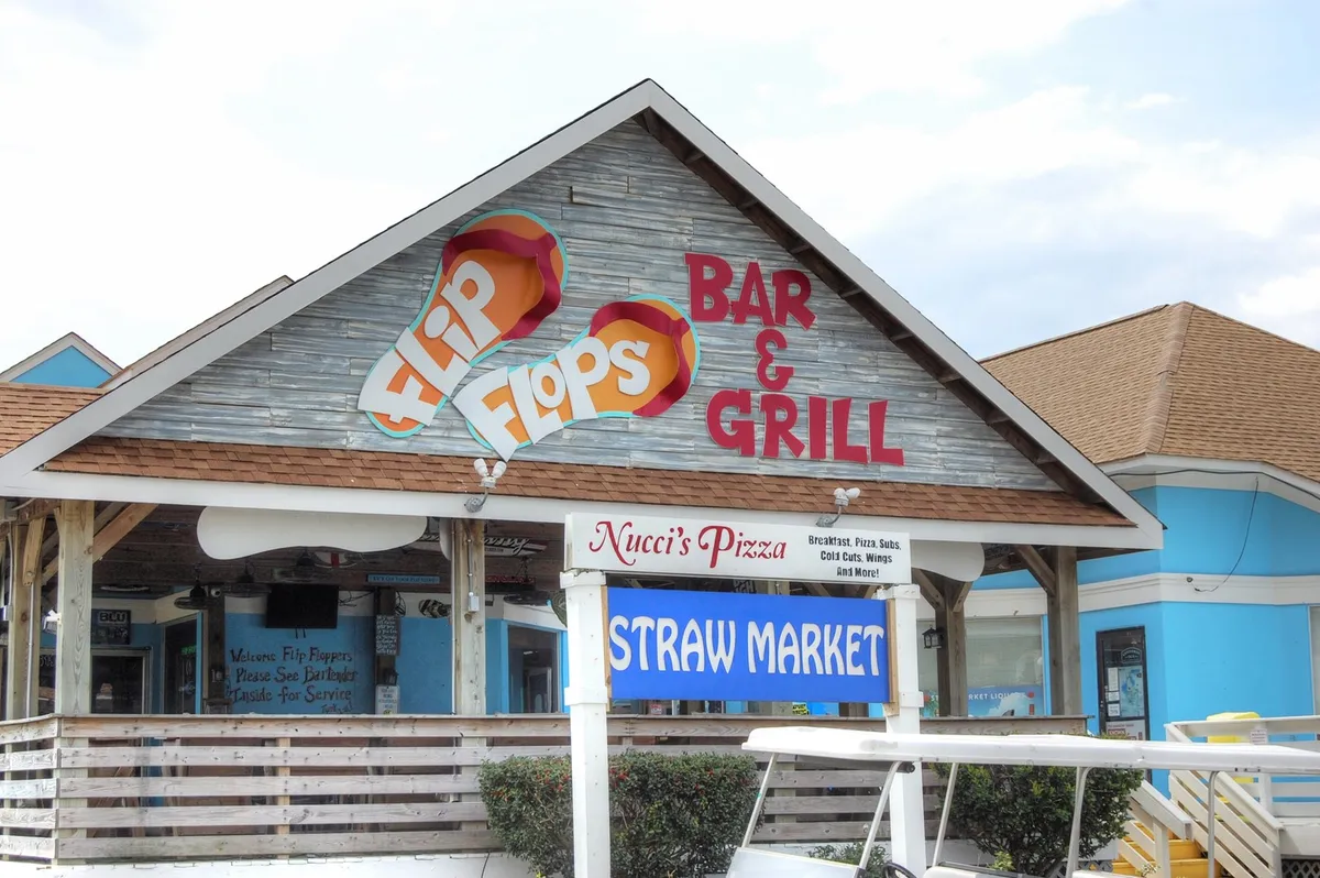 This exterior image captures the lively storefronts of Flip Flops Bar & Grill, Nucci's Pizza, and the Straw Market, set in a relaxed, social atmosphere. People are likely enjoying food, drinks, and shopping, reflecting the accessible amenities near Mariner's Cove.