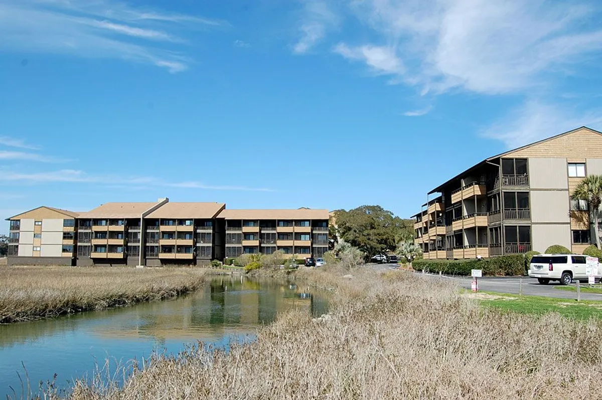 Two multi-story condo buildings sit beside a calm waterway, surrounded by lush vegetation. The setting sun bathes the area in warm light, highlighting the natural beauty and inviting guests to explore the outdoors.