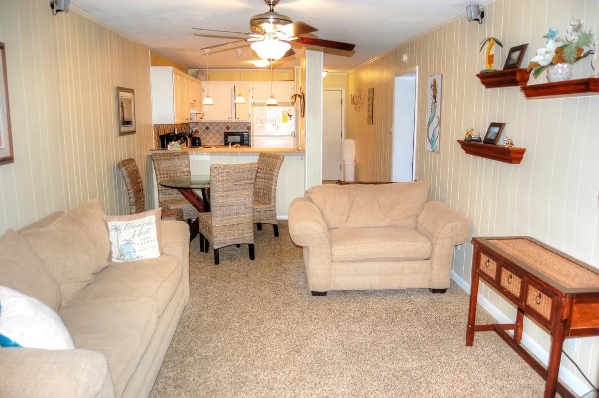 A bright, open-concept interior featuring a comfortable sofa, a dining table set for several people, and a modern kitchen area with decorative shelving. The space is styled with coastal touches, encouraging relaxation and togetherness, and the layout seamlessly flows from living to dining to kitchen—perfect for gatherings and family meals.