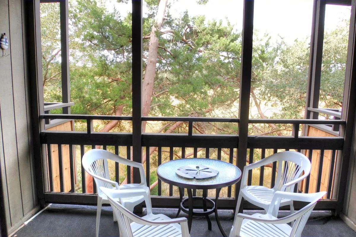 The image shows a tranquil screened-in porch featuring two inviting white chairs and a small round table, with lush greenery visible just beyond the enclosure. The scene suggests a serene space ideal for unwinding, sipping morning coffee, or enjoying quiet conversation, all while being surrounded by nature's calming sights and sounds.