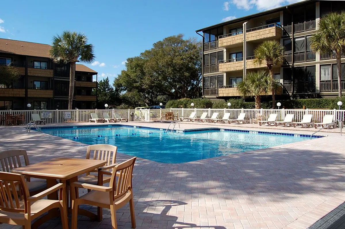 An inviting pool area framed by palm trees and community buildings, with lounge chairs and an outdoor dining table ideal for relaxing after a night out. The setting is perfect for socializing, sunbathing, and enjoying time with friends in a leisurely atmosphere.