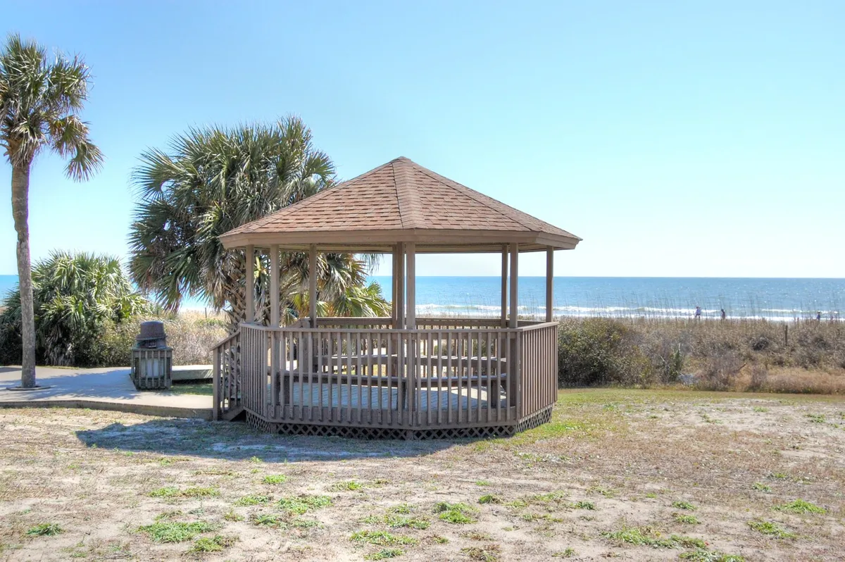 An open wooden gazebo perched near the ocean, surrounded by palm trees and offering sweeping sea views. The tranquil setting suggests a peaceful retreat for quiet reflection, socializing, or simply enjoying the fresh coastal air and sunset vistas.