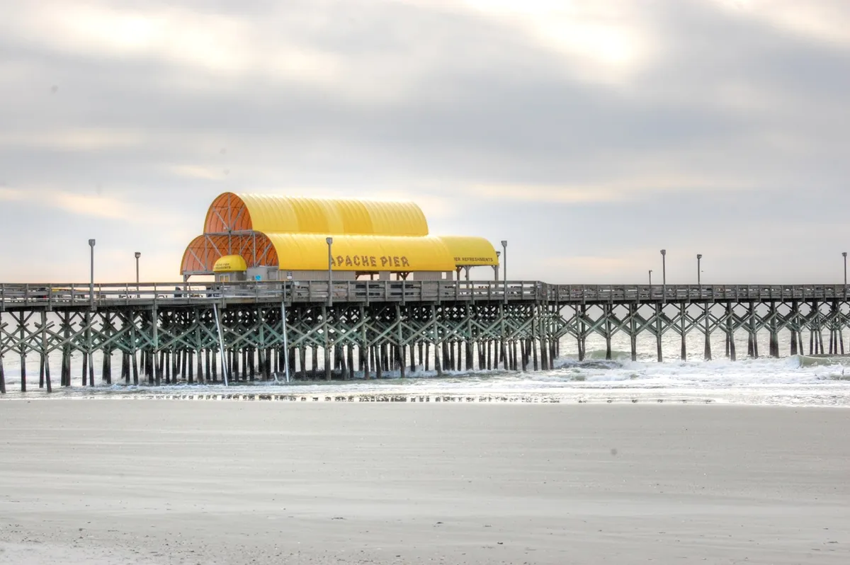 A classic wooden pier with a yellow roof stretches into the ocean, waves rolling below. The early morning light creates a serene ambiance, inviting visitors to take a leisurely walk out over the water.