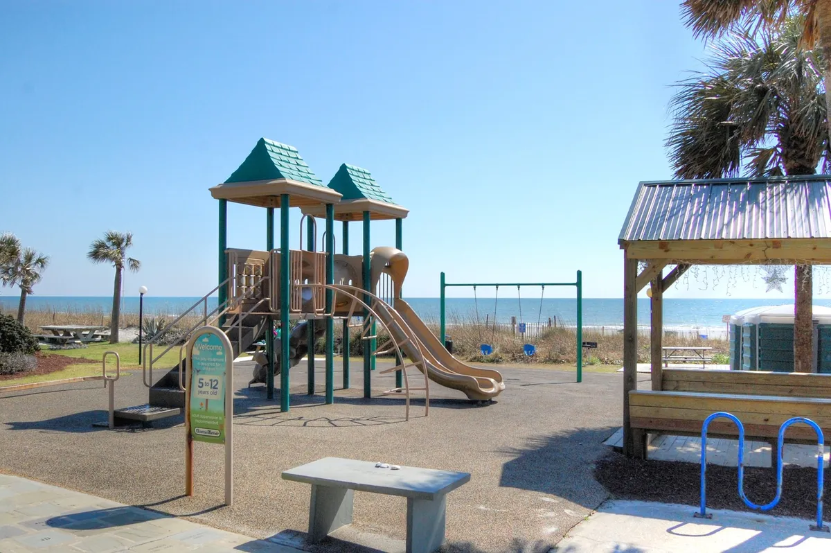 A cheerful beachfront playground with swings, a picnic area, and views of the sandy beach and ocean. Children are playing, while families enjoy picnics and the sea breeze. The sun is shining, creating a relaxed and inviting atmosphere perfect for family fun.