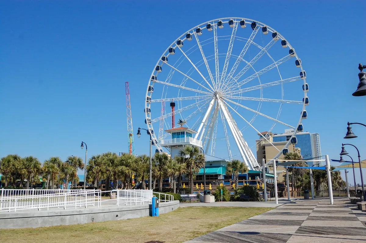 A vibrant exterior shot showcasing the lively Myrtle Beach boardwalk with a Ferris wheel, palm trees, and bustling entertainment venues. Groups of people enjoy rides, outdoor dining, and strolls along the festival-lit boardwalk at sunset, embodying the area's joyous, social energy.