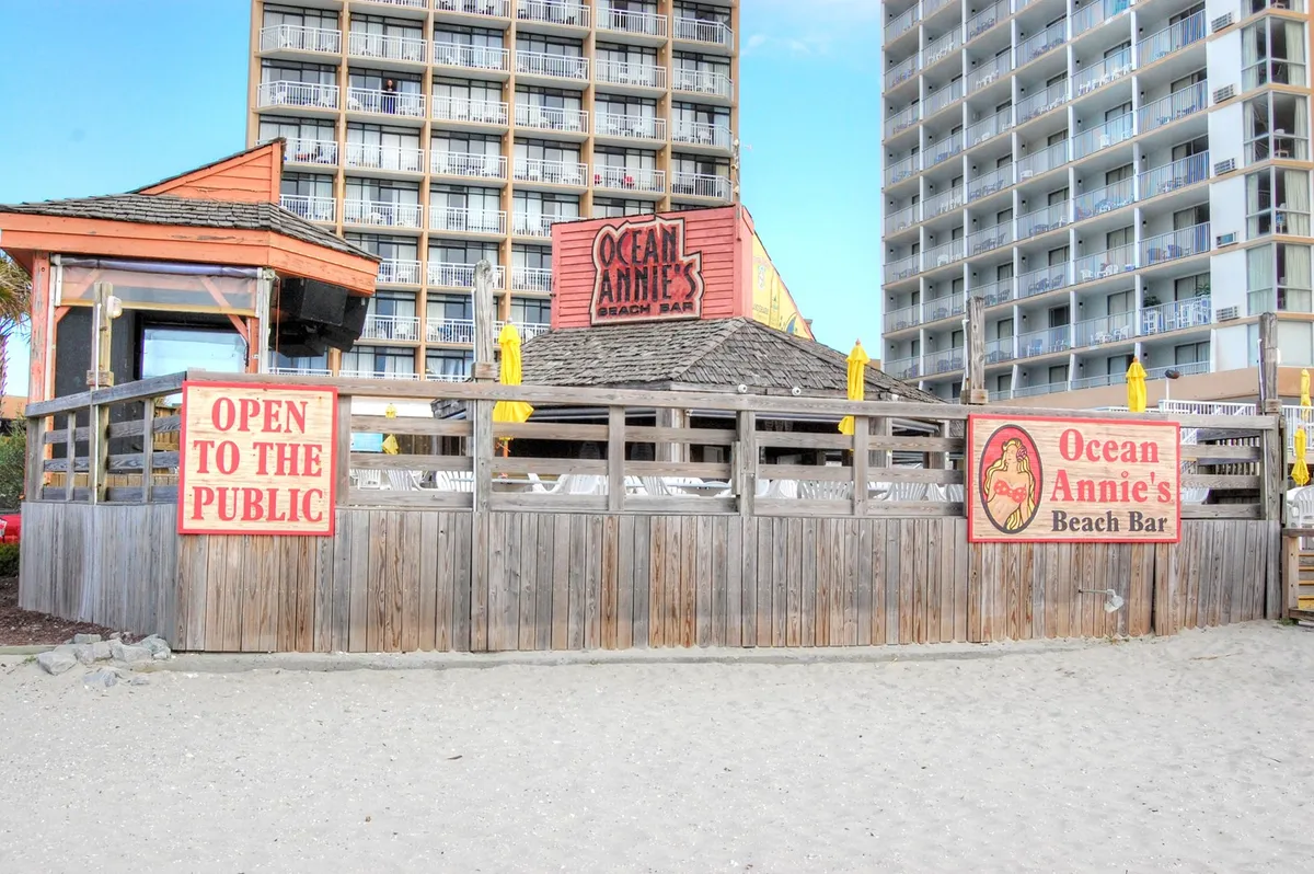 This image captures the iconic Ocean Annie's Beach Bar, with its inviting wooden structure, sandy beachfront, and welcoming signage. The setting exudes a casual, energetic vibe perfect for socializing and live music, with hints of sunset lighting that suggest lively evenings.