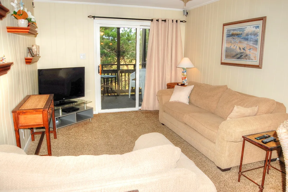 A comfortable living room setup featuring a plush sofa, television, elegant curtains, tasteful artwork, and a coffee table. The ambiance is warm and inviting—ideal for movie nights, reading, or unwinding after a day at the beach.