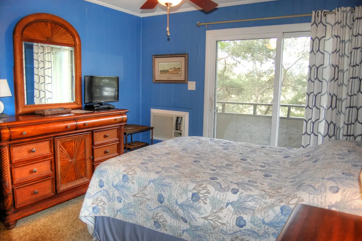 A peaceful bedroom with a floral comforter, wooden dresser with mirror, and sliding glass door opening to a balcony. The room bathes in natural light, providing a serene, private retreat and a seamless connection to the outdoors.