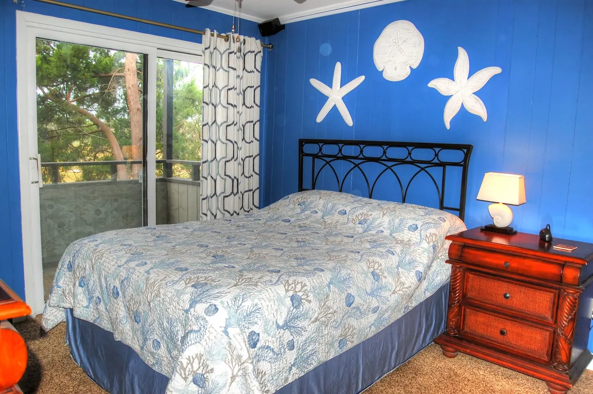 A tranquil bedroom scene featuring a plush bed with blue and white bedding, a decorative starfish on the wall, and a wooden nightstand with a lamp. The soft natural light filtering into the room enhances the serene and relaxing atmosphere, evoking a sense of restful comfort.