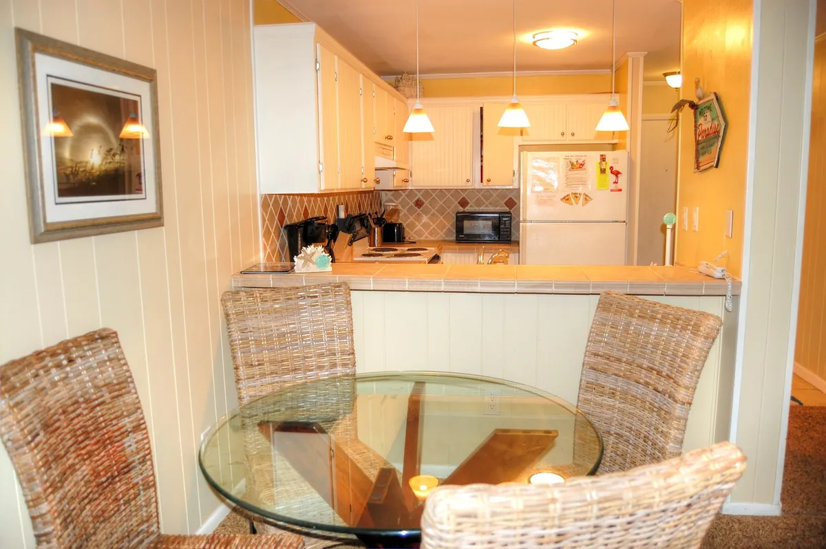 A cozy, sunlit dining area with a glass dining table surrounded by woven chairs, set beside a kitchen with modern appliances and attractive wall art. The space feels intimate yet open, reflecting a relaxed, coastal vibe ideal for both casual breakfasts and group dinners.